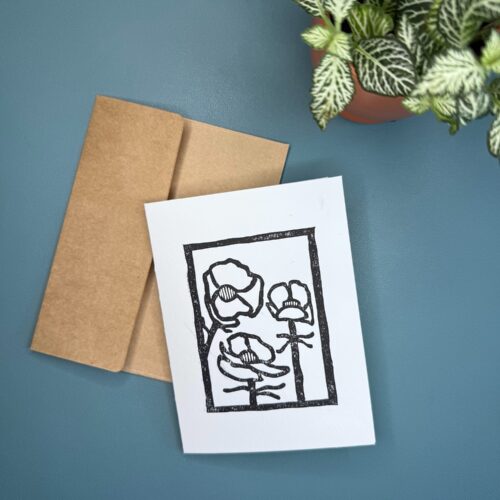 A black ink print of an Alpine Buttercup flower on a white card with a brown envelope on a green background with a plant