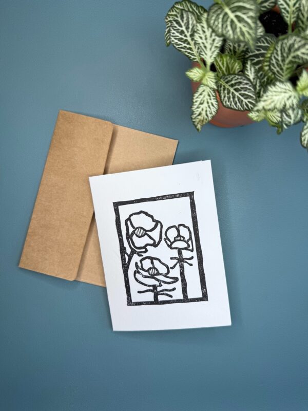 A black ink print of an Alpine Buttercup flower on a white card with a brown envelope on a green background with a plant
