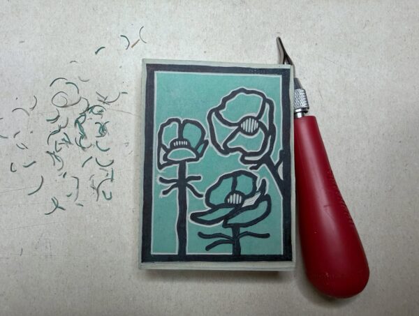 A red carving tool sits beside a partially carved green printing block with a black Alpine Buttercup flower drawn on it on a grey background.