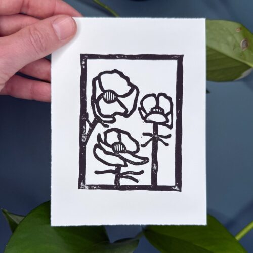 A black ink Alpine Buttercup print on white paper is held in front of a green background with a plant.