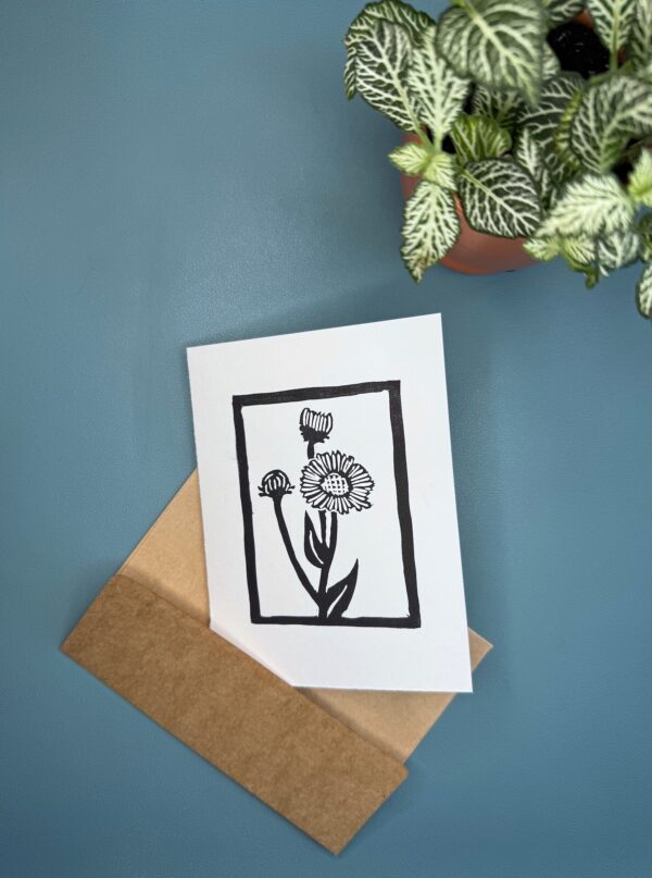 A black ink print of an Aspen Daisy flower on a white card with a brown envelope is sitting on a green background with a plant