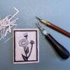 Black and brown carving tools sit beside a partially carved grey printing block with a black Aspen Daisy flower drawn on it on a green background.