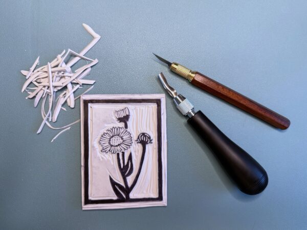 Black and brown carving tools sit beside a partially carved grey printing block with a black Aspen Daisy flower drawn on it on a green background.