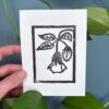 A black ink print of a clammy groundcherry flower on a white card is held by a hand above a green background with a plant