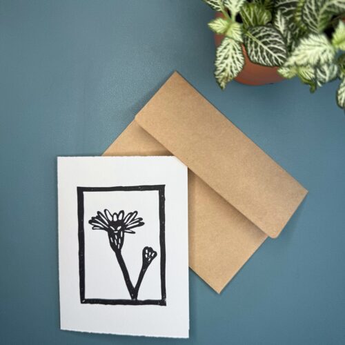 A black in print of a Marsh Aster flower on a white card with a brown envelope on a green background with a plant