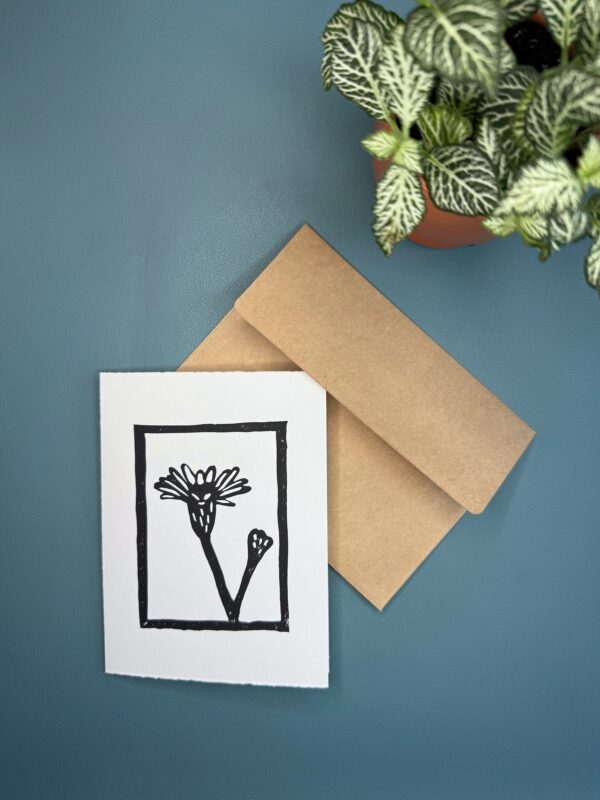 A black in print of a Marsh Aster flower on a white card with a brown envelope on a green background with a plant