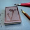 Marsh Aster carving 1 A red and brown carving tool sit beside a partially carved printing block with a Marsh Aster flower drawn on it on a green background.
