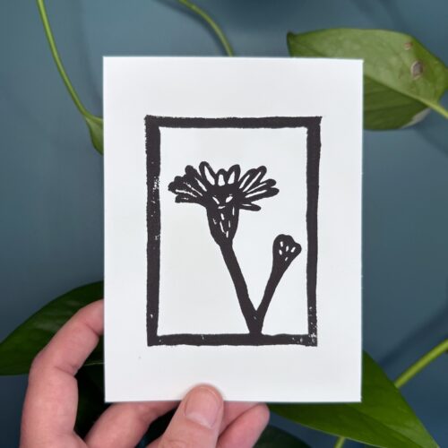 A black ink print of a marsh aster flower on a white card is held by a hand above a green background with a plant