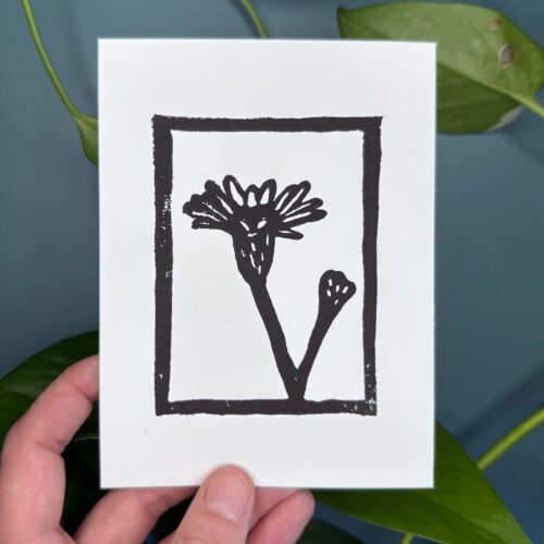 A black ink print of a marsh aster flower on a white card is held by a hand above a green background with a plant