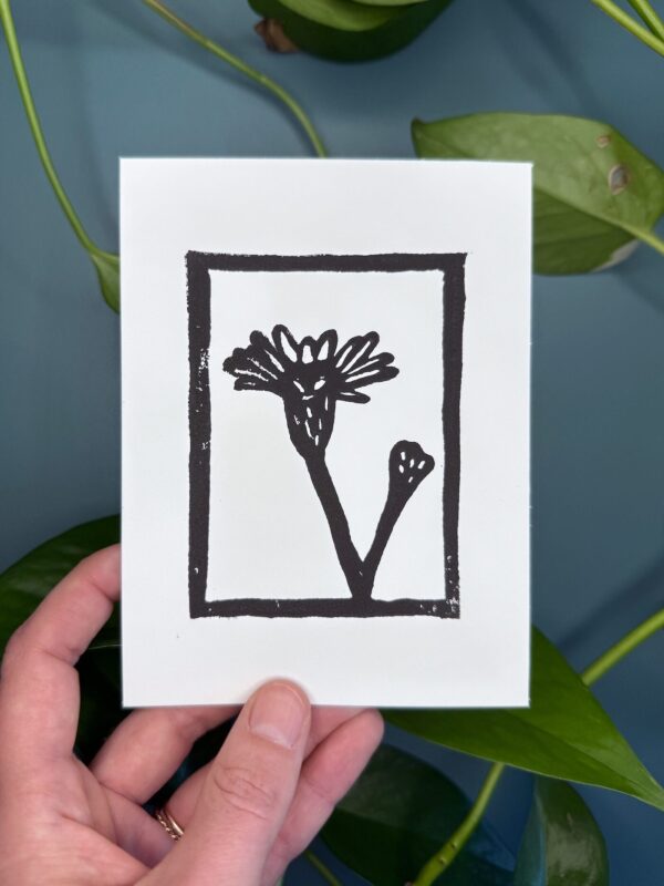 A black ink print of a marsh aster flower on a white card is held by a hand above a green background with a plant