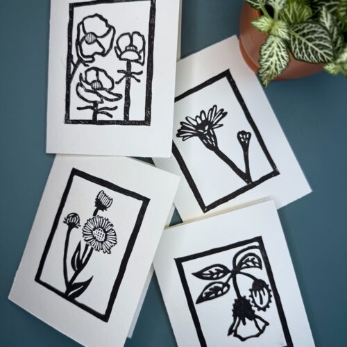 Four black ink wildflower cards on white paper on a green background with a plant