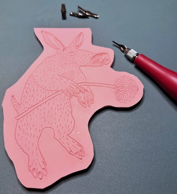 Aardvark progress 2 A red carving tool sits beside a partially carved pink printing block with an Aardvark shape.
