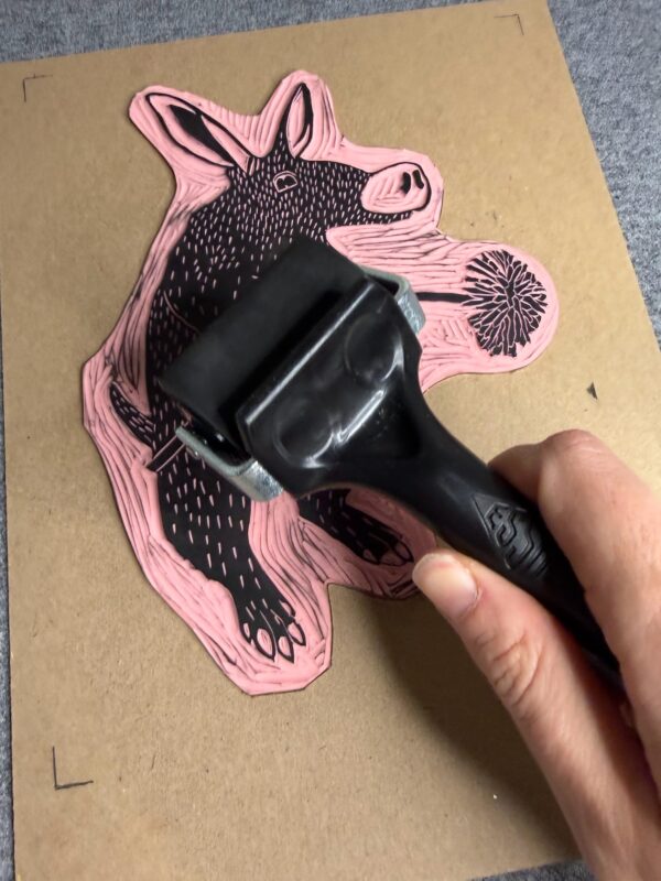 A hand rolls ink with a small brayer onto a pink Aardvark carved block.