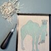 Camel carving A black carving tool sits beside a partially carved grey printing block with a camel shape on a green background.