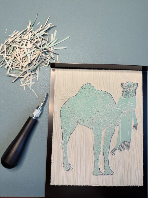 Camel carving A black carving tool sits beside a partially carved grey printing block with a camel shape on a green background.