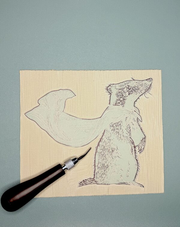 A black carving tool sits beside a partially carved grey printing block with a Badger shape on a green background.